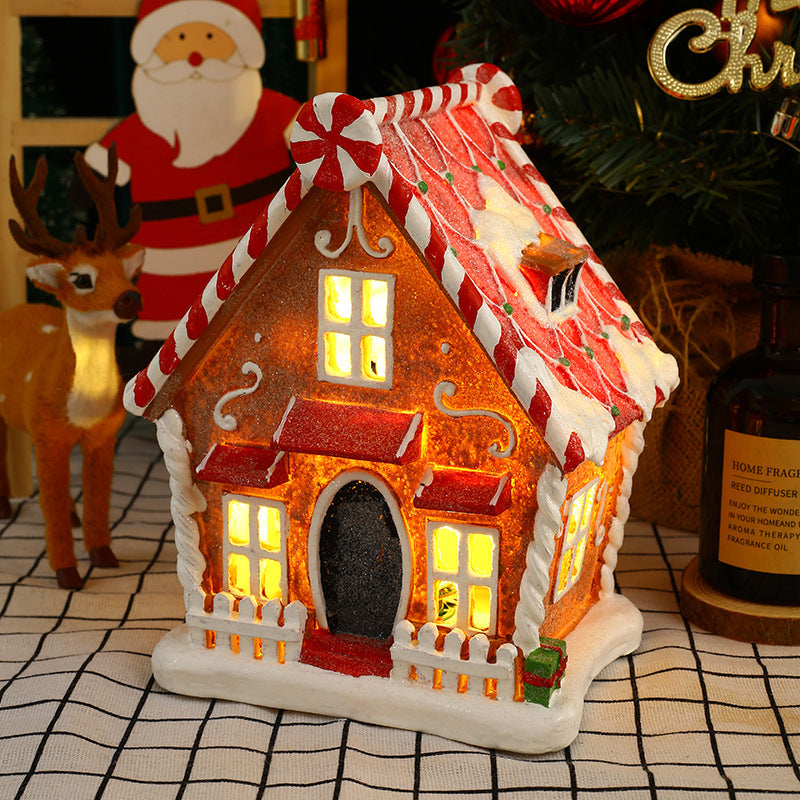 Decorative gingerbread house with lit windows on a checkered surface, surrounded by Christmas-themed items.