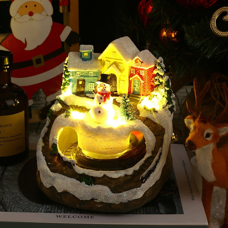 Decorative Christmas scene with a small house, snowman, and reindeer on a lit base.