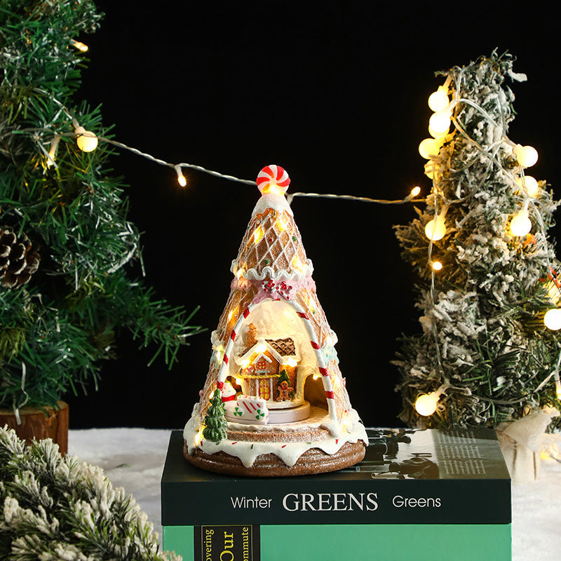 Decorative Christmas tree-shaped gingerbread house with lights, surrounded by festive trees and decorations.